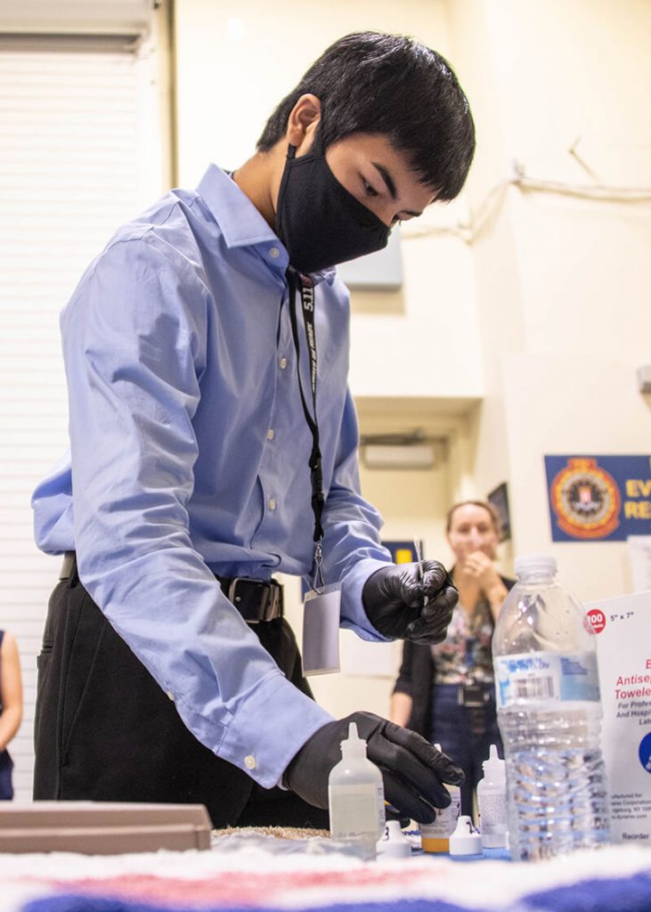 A teenage boy wearing a blue button-down shirt stands at a table wearing black gloves and appearing to work with bottles of liquids.