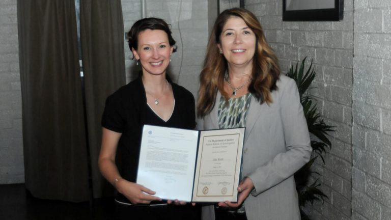 Two caucasian women standing side by side and facing the camera, each holding open part of a certificate encased in a folder.