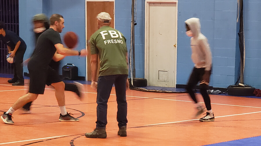 People play basketball in a gym - one man is dribbling the ball, another is wearing a green t-shirt that says FBI Fresno on the back, and a younger person is in motion near them.