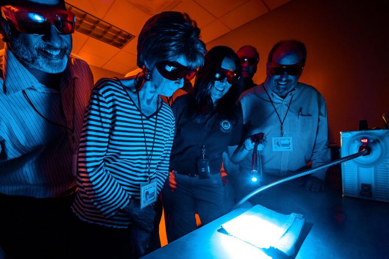 A Special Agent demonstrates using an alternate light source in a dark room. Members are gathered around and behind her, all wearing dark glasses while peering at a glowing blue light.