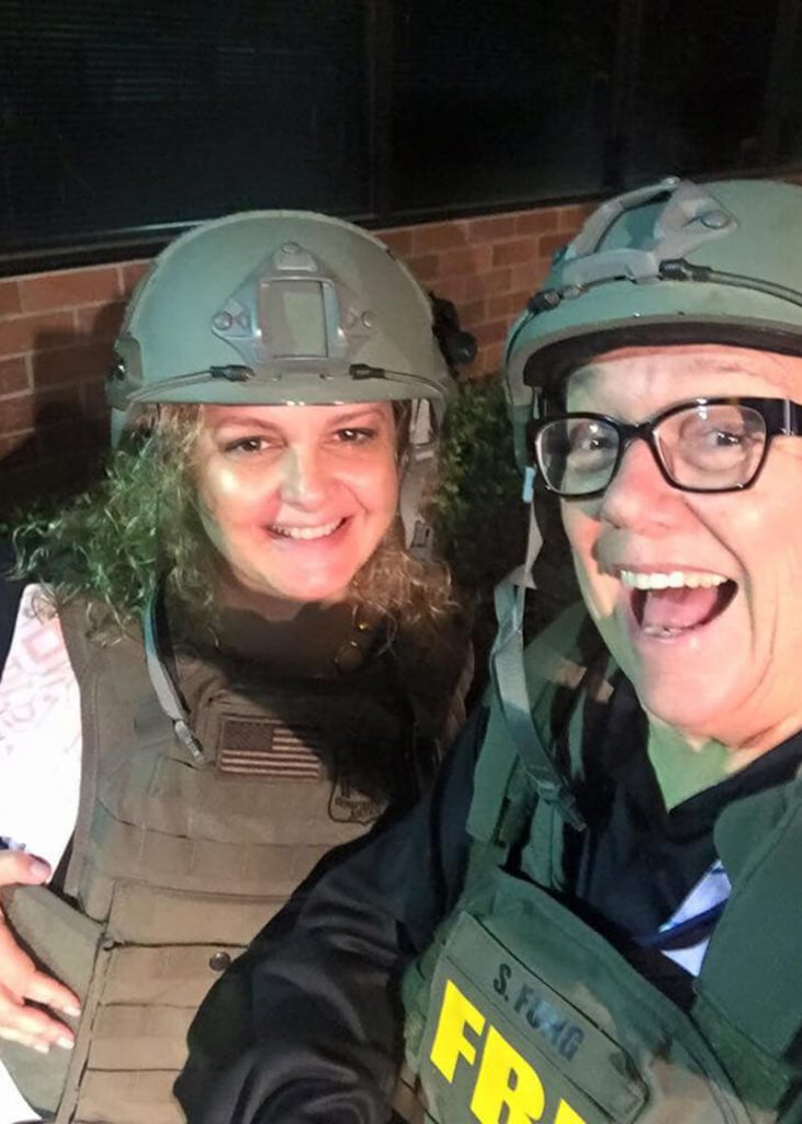 A caucasian man and woman dressed in green combat gear pose for a selfie, smiling at the camera.