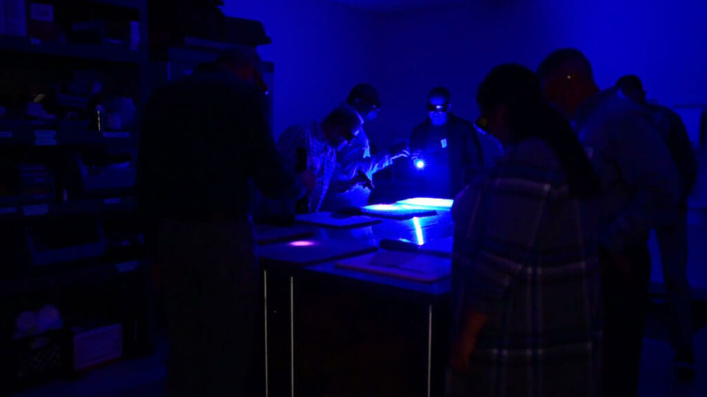 Small group of people standing around a table in the low glow of blue light while they all wear dark glasses and observe an item on the table being illuminated by a man holding type of light.