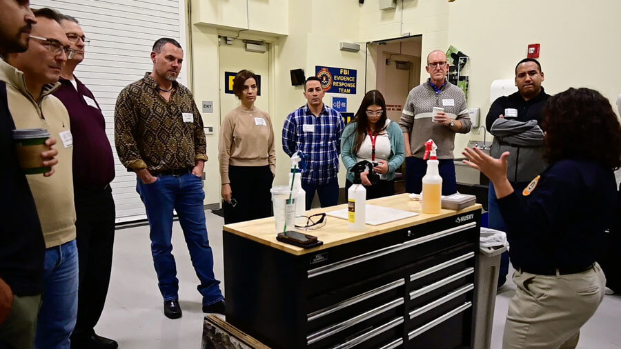 A small group of adults stand gathered around a set of organizational drawers while listening to a person speak and give a demonstration.
